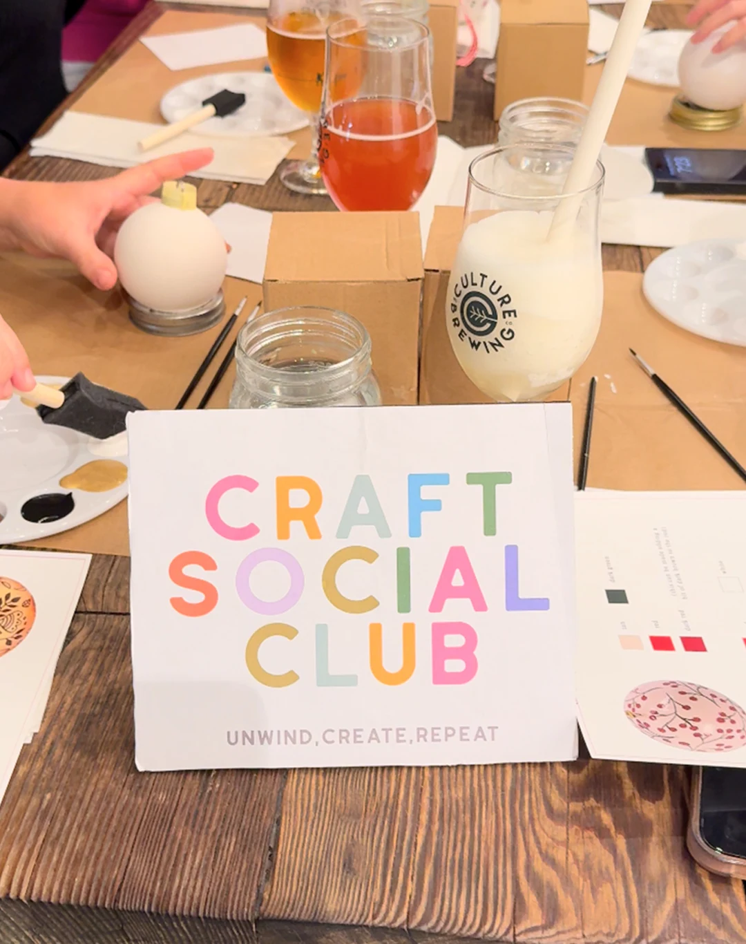Craft Social Club name card at a real workshop table alongside craft supplies and drinks